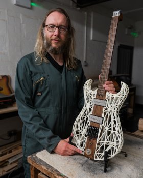 A photo of me stood in the workshop, wearing a set of overalls, with my hybrid wood and 3D printed guitar beside me.