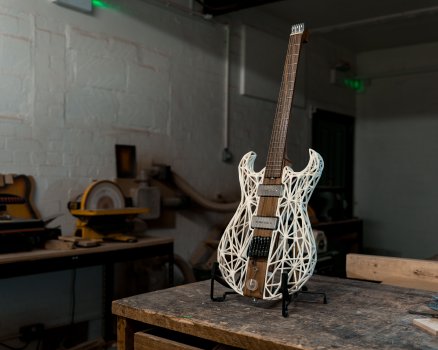 A photo of an unusual looking guitar sat on a stand stood on a workbench in a workshop. The guitar has a central dark wooden core that spans both the neck and the centre of the body, and the body sides are made from a low-polygon white lattice framework that takes the shape of a regular guitar body.