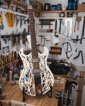 A photo of an  unusual guitar sat on a workbench in the corner of a workshop, with the walls behind the guitar covered with traditional woodworking tools. The guitar itself is an electric guitar that has a central wooden core running the length of the neck and the middle of the body, and then the sides of the body are a 3D-printed white lattice framework.