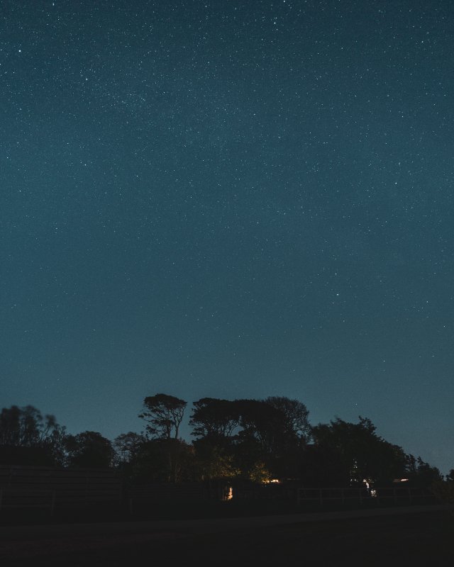 A photo of the night sky over a shiloutted house and trees. The sky is not dark, but you can still make out a lot of stars in the sky thanks to the long exposure.