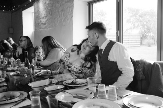 A photo of a couple at a wedding dinner, with other folk eating around them. The bridge has her head on the groom's shoulder with her eyes closed and a gentle smile.