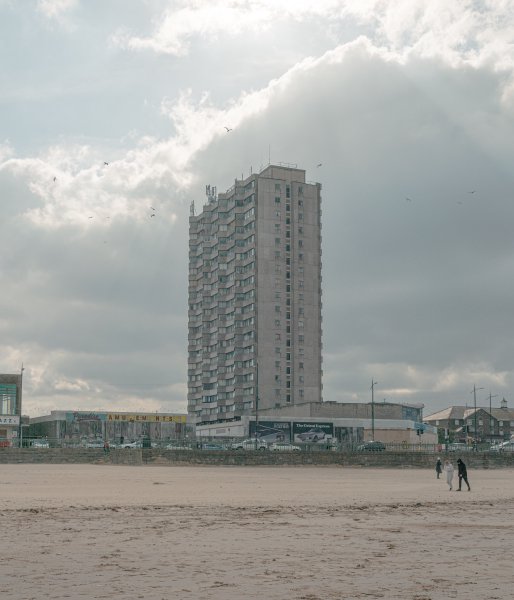 A photograph of a tall brutalistic block of flats that towers alone over the Margate town centre. The photo is taken from the beach, looking back at the prominade, the town, and then the tower.