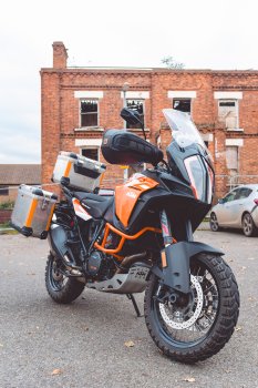 A photo of a big KTM adventure motorbike parked in front of an old red-brick building that has been bruned down inside, and is surrounded with chain link fence. The orange of the bike and the orange look of the brick complement each other.