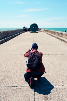 A photo taken behind someone squatted down taking a photograph of a donut looking sculpture at the end of the stone structure we're stood on, with the sea and the sky in the background.