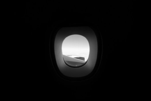 A photo of a window on an airplane. Most of the shot is in total shadow, except the window, through which you can see a blank sky both up and down, and the wing of the aircraft.