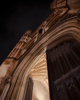 A photo of the front of one of the old college buildings in Cambridge, taken from the ground looking up so we see the gate, the left half of which is open, and then up the detailed carving of the front with its windows and busts. Overhead the sky is black.