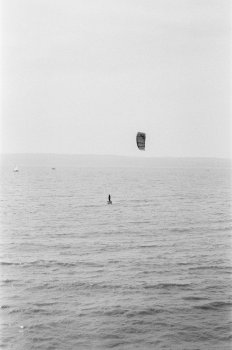 A blown out and somewhat soft photo of the dee estury with wales in the background. In the middle of this frame is a small but contrsting pair of shapes - a surfer on a hydrofoil board and above them a parachutte shaped kite that is pulling them along.