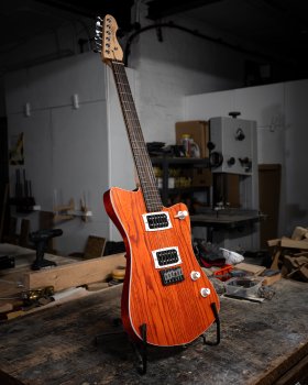 A photo of an orange offset-style electic guitar on a stand on a workbench in a dimly lit workshop. The guitar body has white binding and controls, and two black uncovered humbucker pickups mounted in white mounting rings. The neck is maple with a rosewood fetboard.