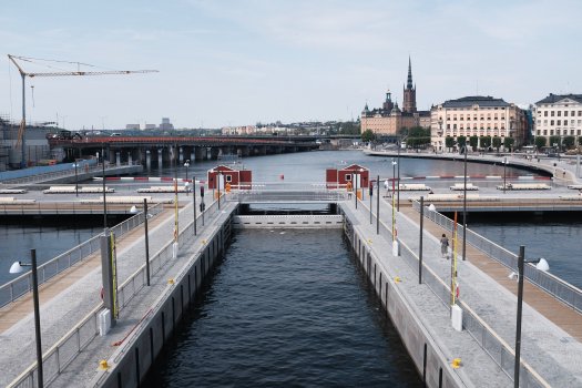 A photo of a set of concrete walls/paths in a body of water in the middle of a city - you can see Stockholm's old town in the background. The photo is over a channel formed between two walls of concrete in the water with paths on, that go to a gate in the middle of the water. On each side at the end is what looks like a red hut in the typical Swedish style, but is in fact covering the mechanism for the gate.
