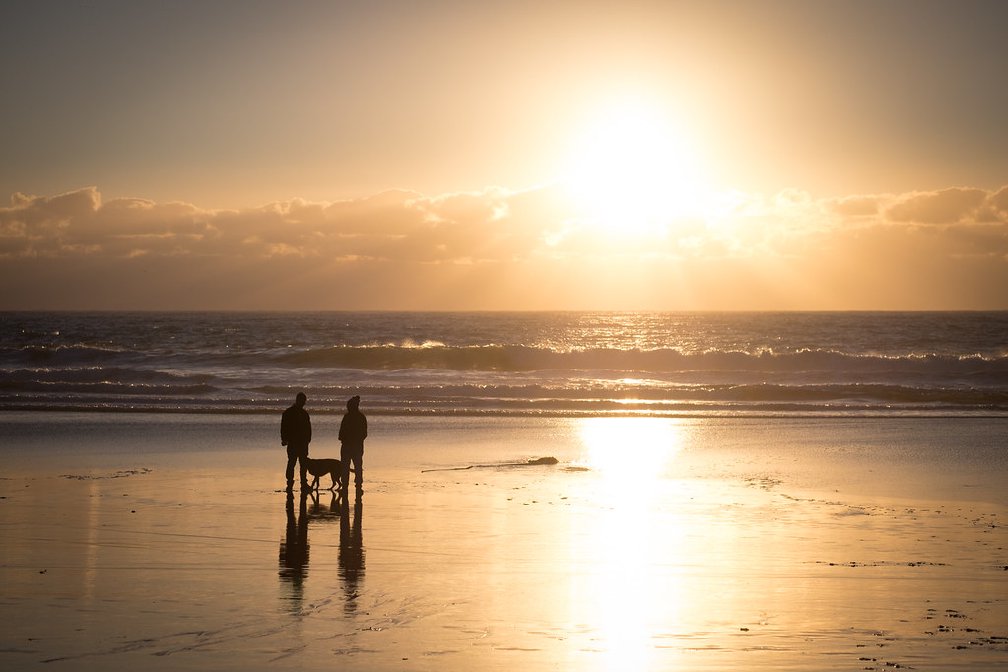 A photo of two people on a beach chatting, with a dog between them. They are on a beach with the sun setting behind them.