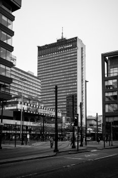 A photograph of a city centre scene, with one tall office block in the center of the frame, which has a sign on the top saying "CITY TOWER"