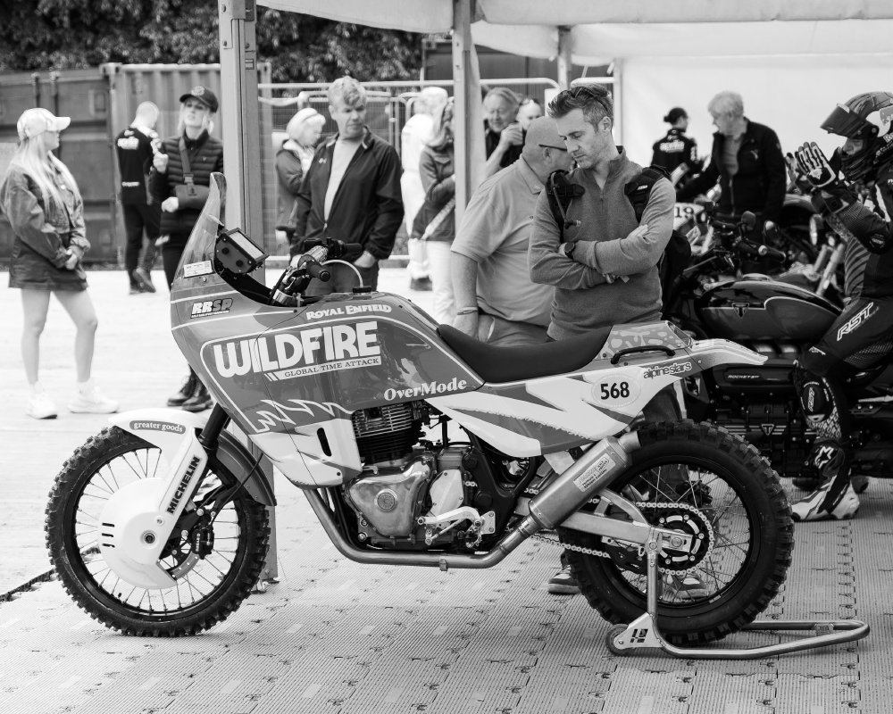 A photo of a man stood amidst the bustle of a motorshow, behind a Royal Enfield adventure style motorbike, his head looking at the bike but with his eyes closed and a calm look on his face.