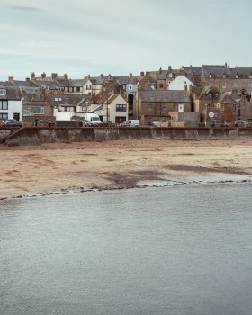 A photo of the town of Eyemouth, taken from the harbour across the bay. In the foreground is the sea, then midway up the frame you have the beach, and above it the concrete wall up to the promenade. Then there is a wall of brick houses, about three rows deep as they go up the hill, with a mix of red brick and white/blue walls. Above that is a slighly overcast sky.