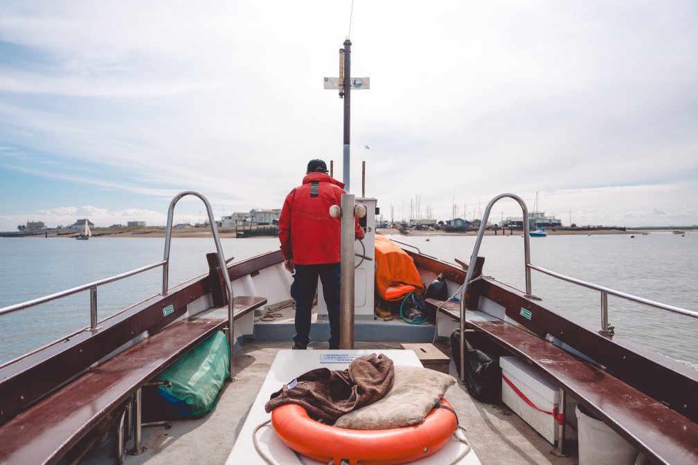 A photo taken from the back of a small boat, looking forward at the pilot driving the boat towards a coast line of buildings that is not that far away.