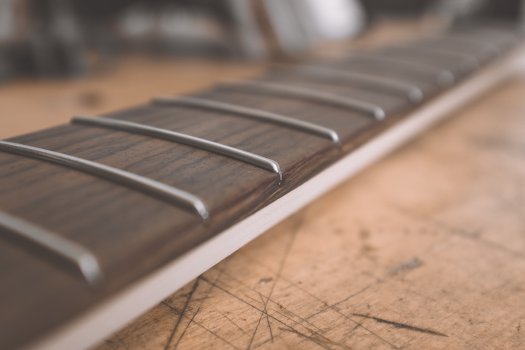 A close-up photo of an unfinished guitar neck on a workbench, showing the detail on the end of a single fret, with the rest being out of focus. The fret end is more rounded than that you might find on a regular guitar.