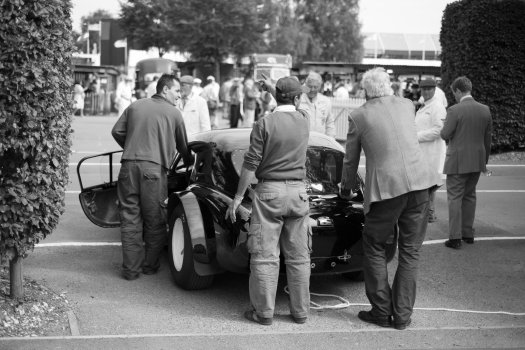 Three men have paused as they push a classic race car, with one pointing in a possible direction they should be going.