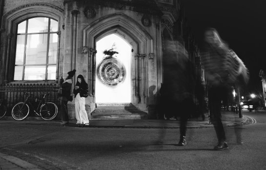 A long-exposure photo of a corner of a building taken at night, on which there is a blurry, overlit clock. To the right of the clock is a couple, blurred as they walk towards us, the main looking at the camera. To the left of the clock are a couple of probable tourists, with one taking a photo with their phone of something beyond the left of the shot, and their friend staring intently at their phone, the glow of which is lighting their face.
