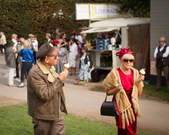 A man and an elderly lady enjoying an ice-cream on a sunny day. The man is dressed as a US WWII army pilot, and the lady in furs from a similar period.