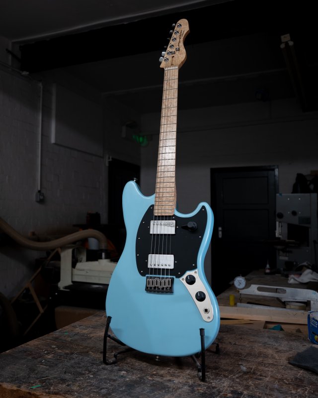 A photo of a blue painted offset style guitar stood in a stand on a workbench in a moodily lit workshop. the guitar has black pickguard with two chrome covered humbuckers in it, and has a birds-eye maple neck and fretboard.