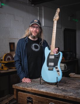 A photo of me stood next to a guitar in a stand on a workbench in a moodily lit workshop. The guitar is a pale blue offset electric guitar with a black pickguard and two chrome covered humbucker pickups, along with a birds-eye maple neck. I'm stood with one hand on the guitar, looking into the camera and smiling.