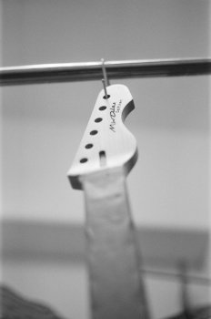 A film photo of a guitar neck hung up on a rack, looking upward from the ground. The fretboard is taped up. Most of the photo is in soft focus except the headstock, which has my signature on it and then the name of the guitar "DELFINEN"