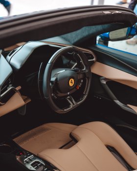 A close-up photo showing the steering wheel and drivers seat of a modern Ferrari car. The steering wheel is black with light coloured stitching on the inner rim. It has a number of buttons and switches on it and carbon-fibre paddle shift leavers, and in the middle is the bright yellow circle with the black prancing horse logo on it.