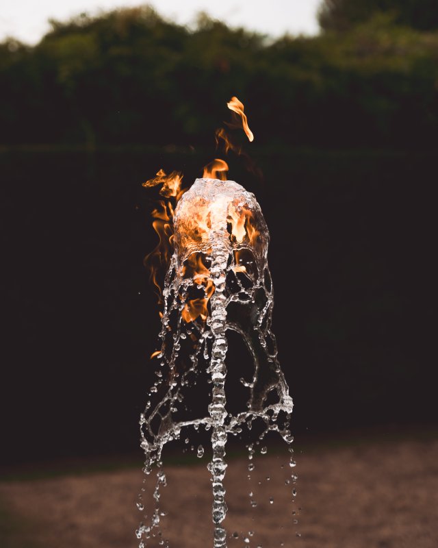 A photo capturing the top of a water fountain, freezing it in time so you can see all the droplets, and inside the top of the fountain as it falls back on itself is fire, with some flames licking off above.