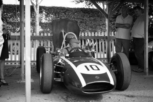 An elderly man is at in the cockpit of a vintage formula one car as the car is prepped.