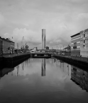 A view along a section of canal in a city center to a bridge about 100m away, with roads on either side. Over the bridge, and mirrored in the water, is a large tower block, that is squarish, but looks like it twists in the middle.