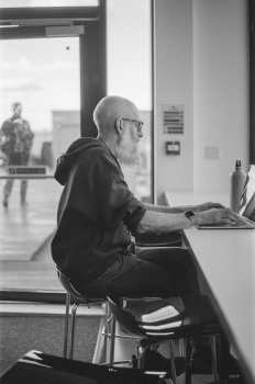 A photo from side on with Jonathan sat on a stool at a bench working on his laptop intently.