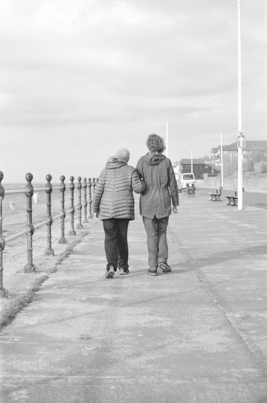 A grainy black and white film photograph of two women walking along a prom away from the viewer, one talled on younger than the other. They walk arm in arm into the distance.