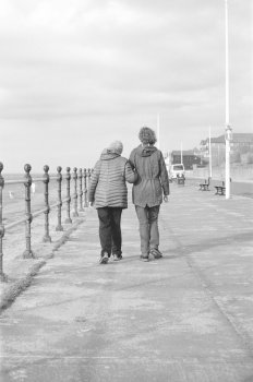 A grainy black and white film photograph of two women walking along a prom away from the viewer, one talled on younger than the other. They walk arm in arm into the distance.