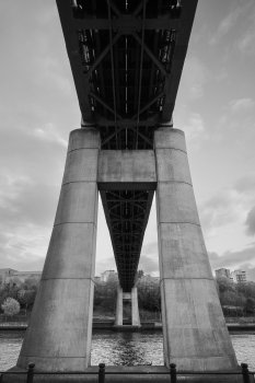 A photo taken under a bridge with a wide angle lens, so you can see the ground under the bridge in front of you, a river, the legs of the bridge and the bridge going over you all the way to the top of the frame.