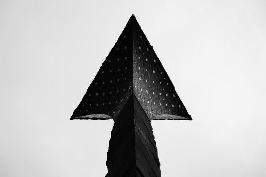 A photo looking up at a triangular top section of sculpture, a bit like a flower in how it expands out from the column supporting it. The sculture is in shadow against a featureless light grey sky.