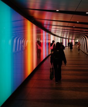 A photo taken down a walkway tunner, where the left wall is made from panels of light in different rainbow colors. People walk down the tunnel, mostly dressed in black.