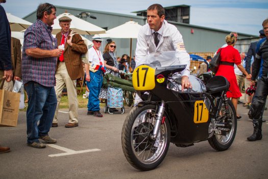 A man in vintage garage outfit pushes a classic race motorcycle through the paddock with a look of determination on his face, surrounded by a mix of people dressed in modern and vintage clothes.