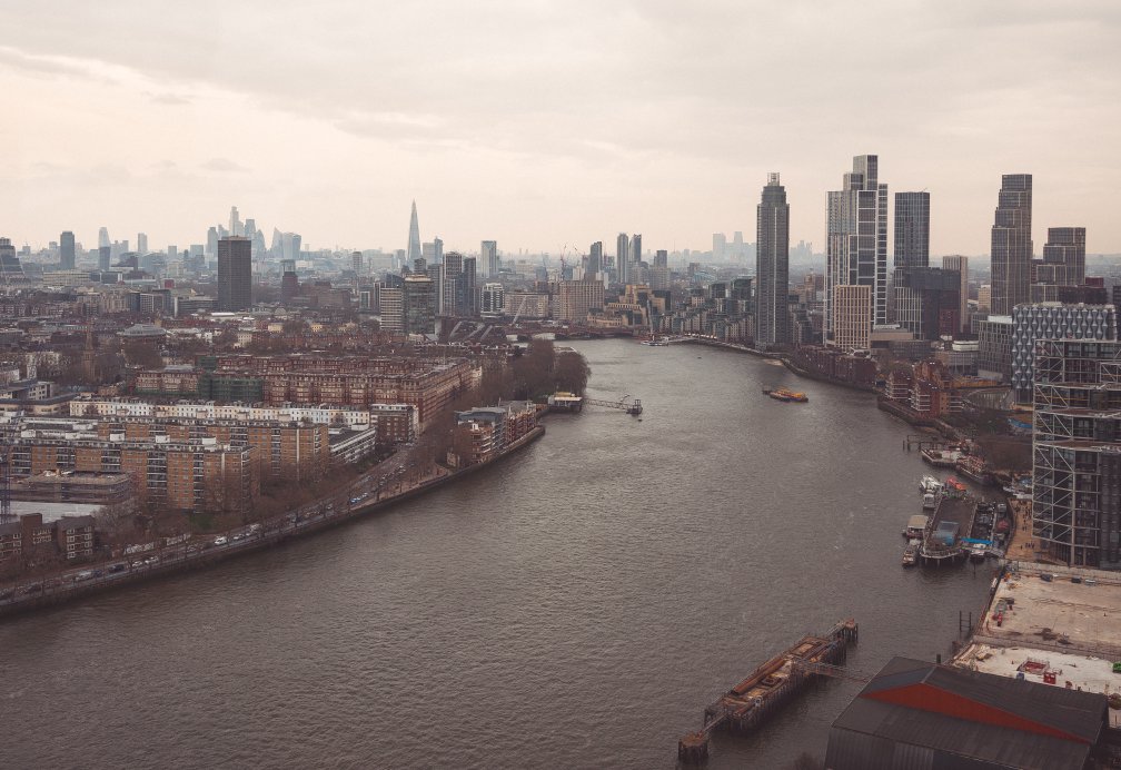 A photo looking down the sweep of a curve in the River Thames as it heads into central London. The sky is overcast, and below that you can see the London sklyine with many tall buildings. In the foreground you look down to see the river sweep past Vauxhall on the right and Chelsea on the left, the latter being full of buildsings made from brick.