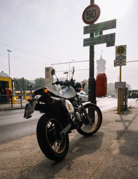 A photo of an old trails bike parted on the sidewalk next to a street lamp covered in road signs take in central Berlin. The sun is high in the sky behind the bike, leaving a lot of it in shadow, but the tank is pale blue, and if you were to look closely you'd see a "Rockstar games" logo stuck to it also.