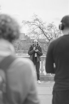 A black and white film photo of a man speaking into a microphone as spotted between the shoulders of two onlookers.