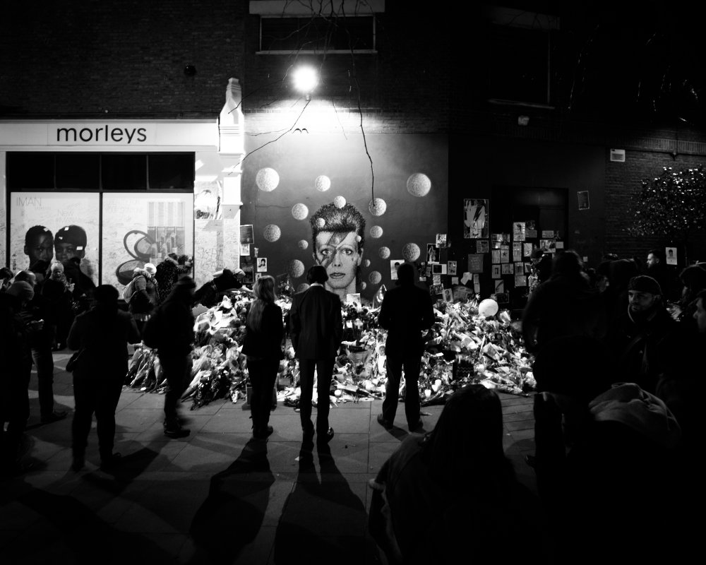 A mural on a wall showing David Bowie as Ziggy Stardust, surrounded with both flowers and mourners looking on in silence.