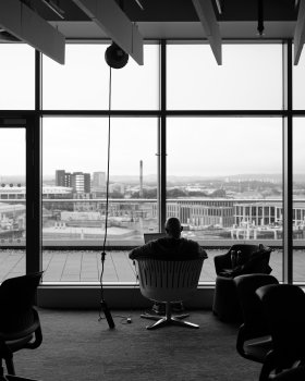 A photo taken of a man sat in a chair with his back to us, working on a laptop. Before him is a floor to ceiling window that looks out over a panoramic view of the south side of Glasgow, this being the 7th floor of the building we're in.