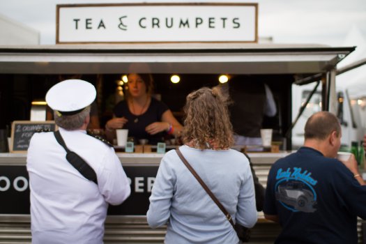 Laura stands in a queue with other people in vintage dress at a van with a sign saying "Tea and Crumpets"