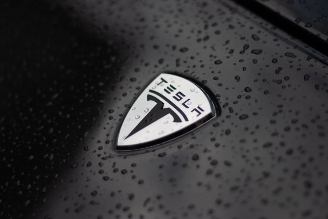 A close up of the emblem of on Telsa car, covered in rain drops.