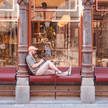 A photo of a man sat on a bench outside a cafe, sat side on to the cafe window with his legs along the bench. He's holding an iPad and a stylus working on something. In the refection in the window you can make out a blurry image of a photographer taking the picture.