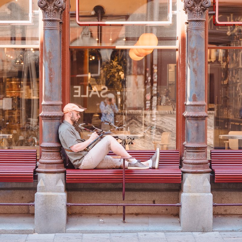 A photo of a man sat on a bench outside a cafe, sat side on to the cafe window with his legs along the bench. He's holding an iPad and a stylus working on something. In the refection in the window you can make out a blurry image of a photographer taking the picture.