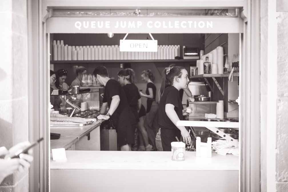 A view through the serving hatch at an ice cream parlour from out in the street, showing all the young workers filling the orders on a sunny day. The sign above the hatch says "QUEUE JUMP COLLECTION" and "OPEN", and off to the left you just can see a phone in someones hands, probably ordering their ice cream.