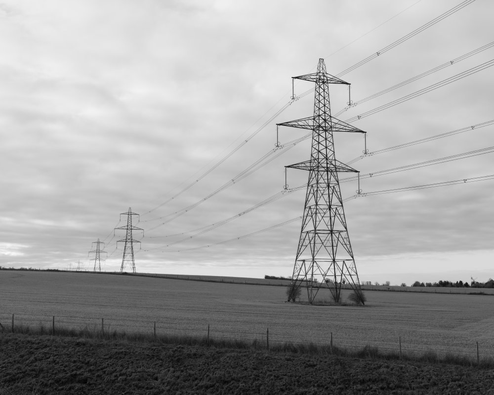 A black and white photo of some rolling fields, and then in the foreground is a large power pylon, part of a line of such pylons that you can see stretch in the distance.