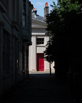 A photo of a white building with a red door caught in the bright sun, with the photo taken from down an alleyway of houses on the left and trees in the right, all of which is almost black in dark shadow, making the door pop out visually.