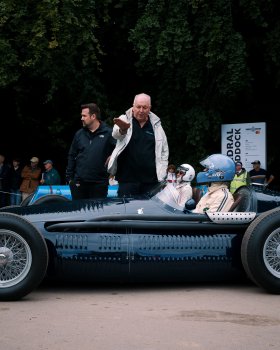 A photo of a classic cigar shaped formular one car on the road, with the driver looking at an older man stood next to the car talking, gesturing with his hand a sort of right hand turn.
