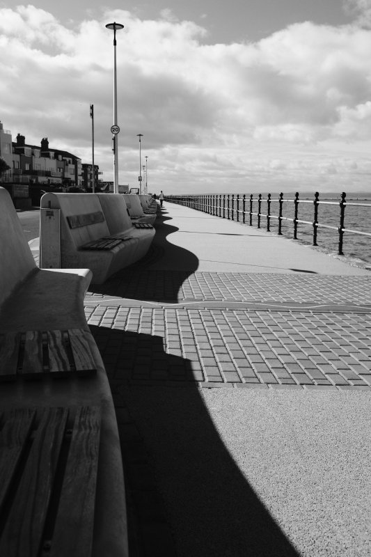 A view along a promenade along the sea front: sea to the right, houses to the left, walk way going into the distance in the middle. Along the left hand edge of the walkway/promade is a wall of concrete benches that are curved back and forth over the course of a few meters interval, and the shadow cast by the sun behing behind them looks like a line of waves.